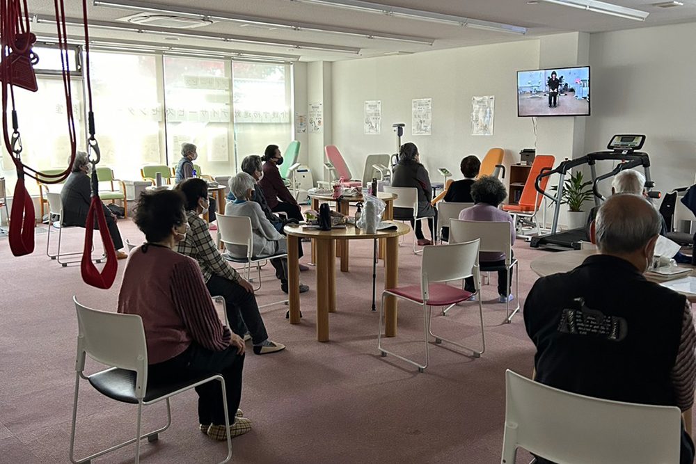 デイサービスセンター めぐみ 御室の広々としたトレーニングルーム。利用者の方々が椅子に座り、正面のモニターに映し出されるリハビリ動画に合わせて、一斉に機能訓練を行っている様子。