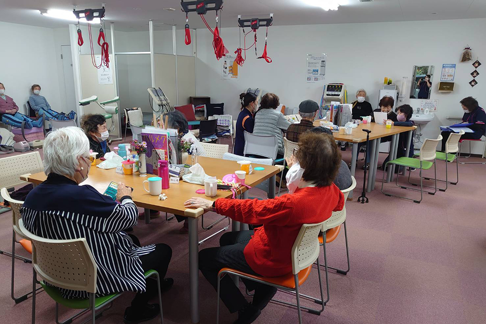 明るく開放的なホールで過ごす利用者の方々の様子。大きなテーブルを囲んで談笑や読書を楽しみながら、天井から吊り下げられたレッドコードなどの訓練機器も備わっている充実した施設内部。